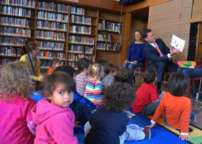 Photo: group of kids sits in a library while man reads to them.